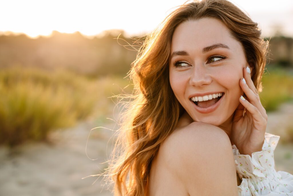 A woman smiles joyfully while seated on the beach, with the ocean waves gently lapping at the shore in the background.